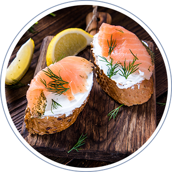 Primer plano de dos tostadas rústicas coronadas con delicadas lonchas de salmón ahumado de Humos de Panitao y una porción de queso crema, decoradas con eneldo y servidas con un trozo de limón.